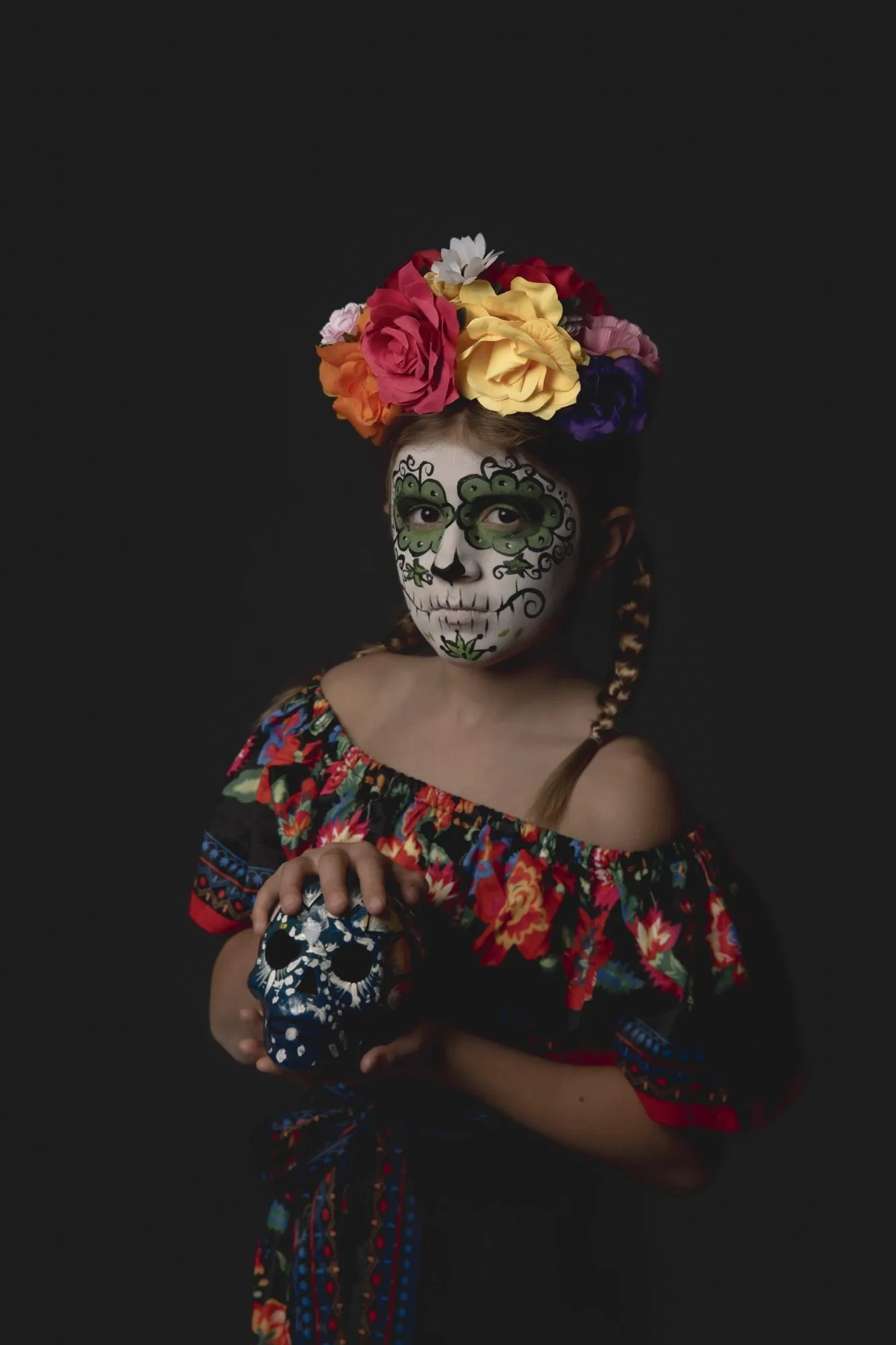 Girl in green sugar skull makeup and a black floral dress holding a painted skull against a dark studio backdrop