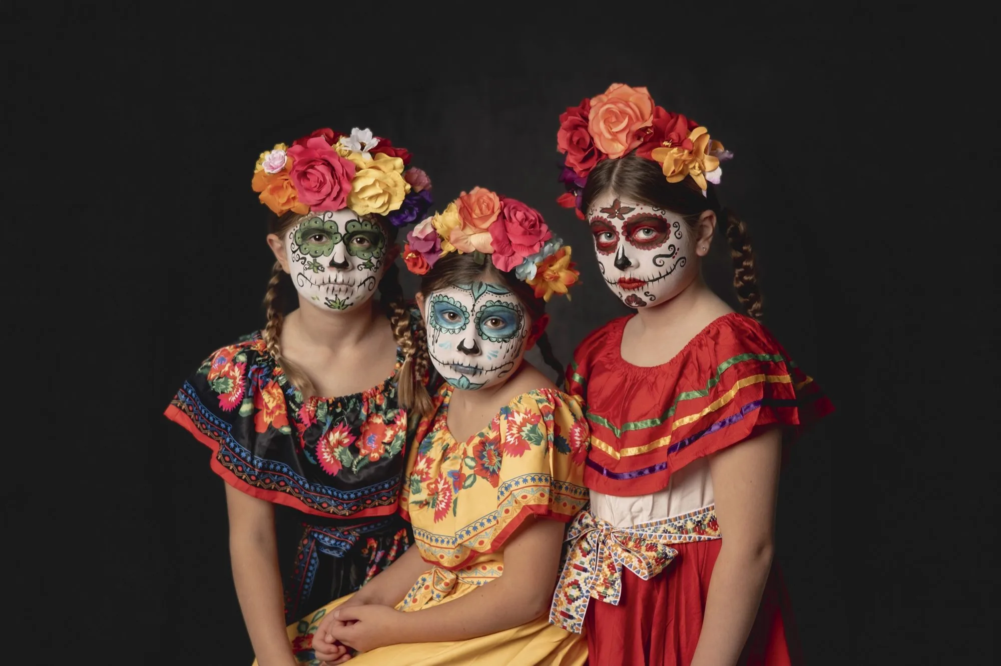 Three girls in colorful Halloween costumes and flower crowns seated together against a black backdrop