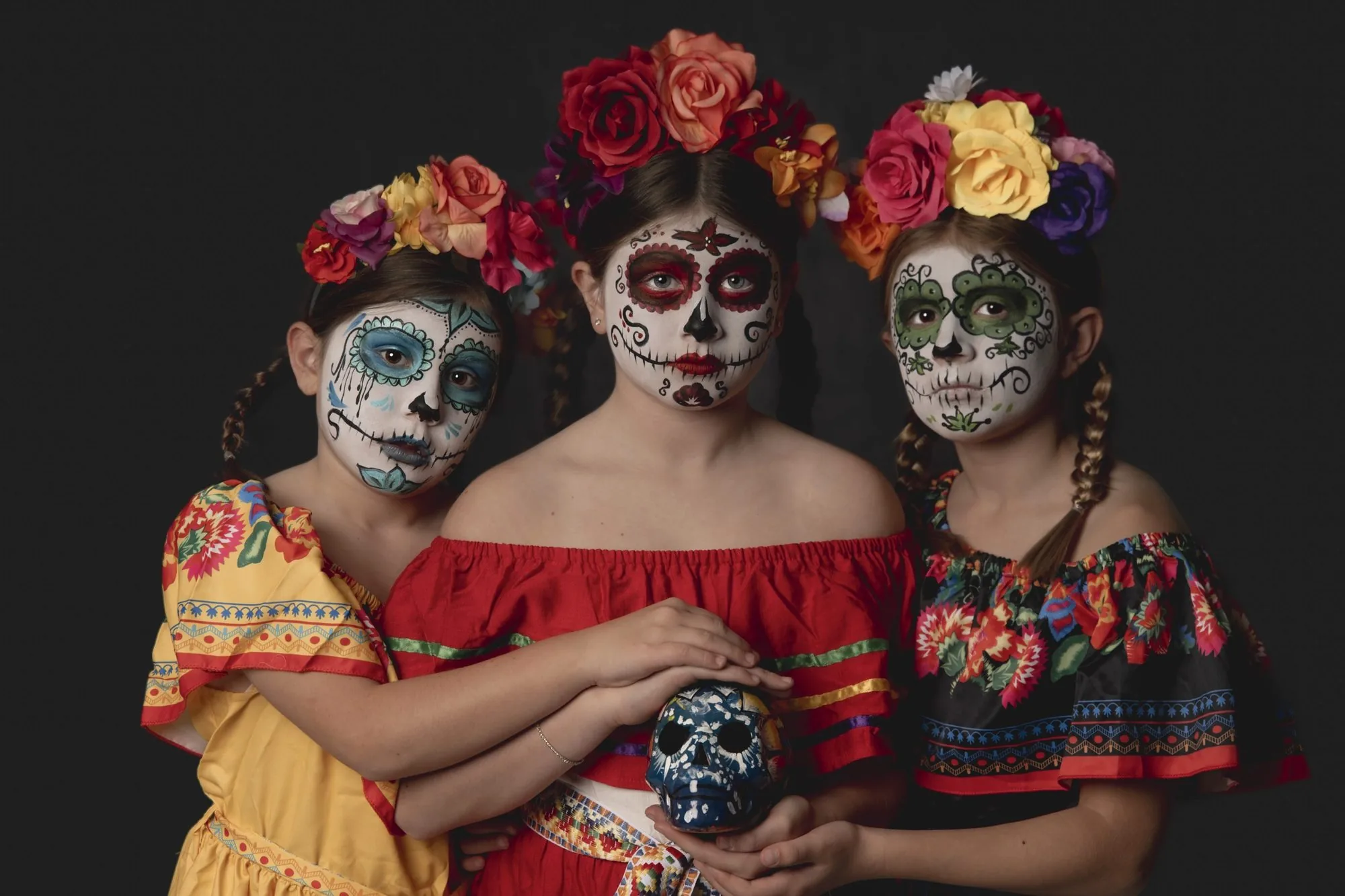 Three girls in sugar skull costumes holding a hand-painted skull together in a studio portrait